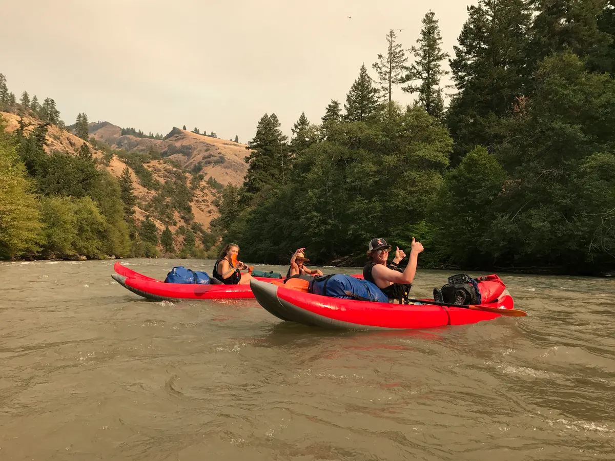 Two loaded inflatable kayaks on a silty river between dry hills and pines, Owen in the near boat giving a thumbs up