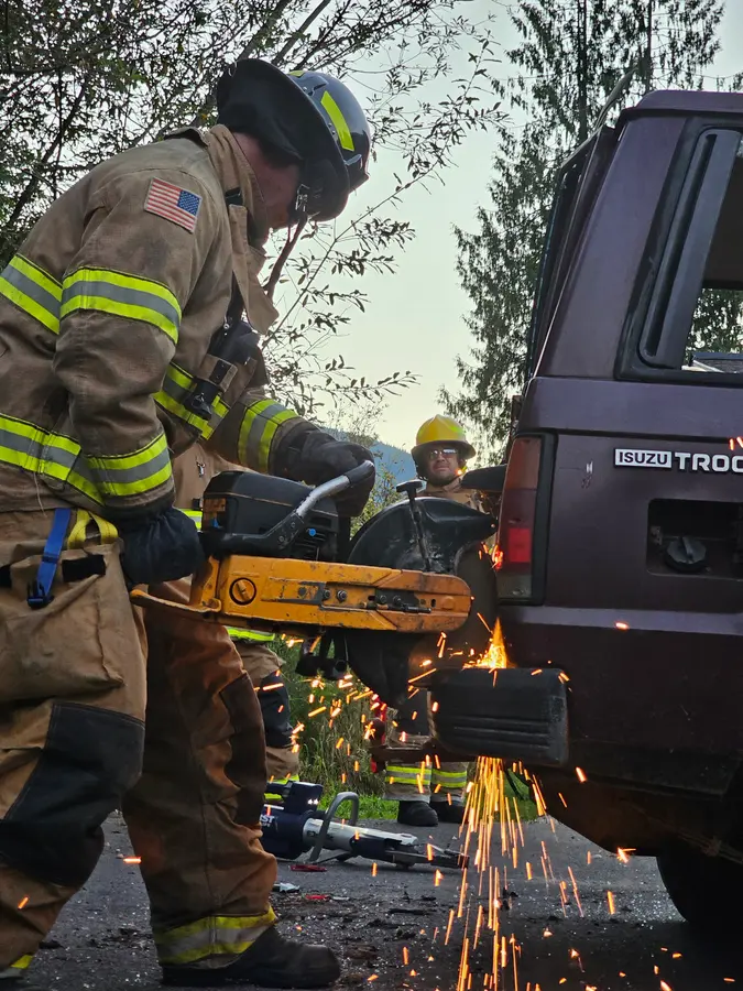 Owen in turnouts running a rotary saw into the rear of an Isuzu Trooper, sparks flying, another firefighter watching