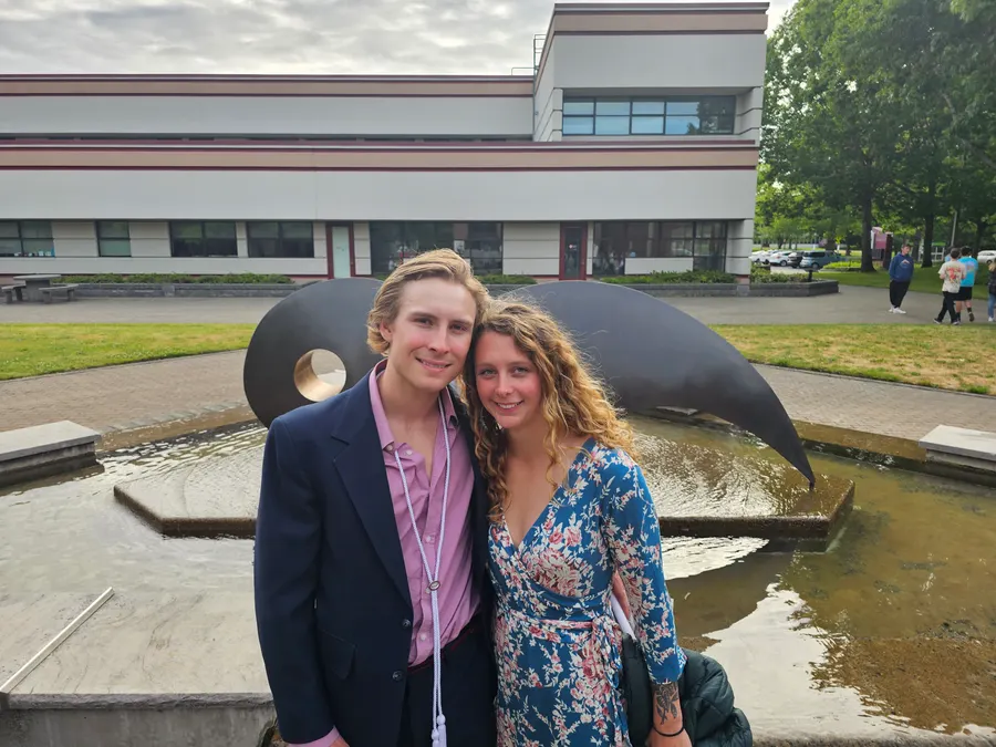 Owen in a navy blazer with white honor cords, standing with his partner in front of the fountain at Whatcom Community College